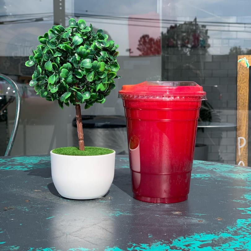 A vibrant red juice in a clear cup sits next to a small potted plant on a distressed table.