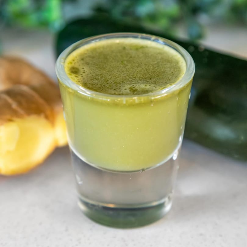 A vibrant green juice shot in a glass, with ginger and cucumber in the background.