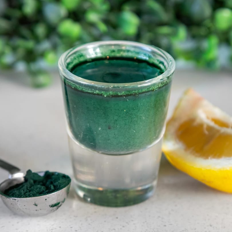 A shot glass of dark green juice, a spoonful of green powder, and a lemon wedge.