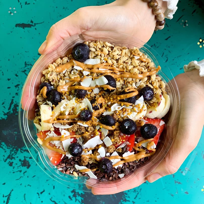 Hands holding a Juice Parlor NoHo acai bowl with granola, blueberries, strawberries, bananas, and peanut butter drizzle.