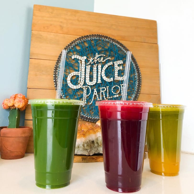 Three colorful juices in clear cups, with a "The Juice Parlor" string art sign in the background.
