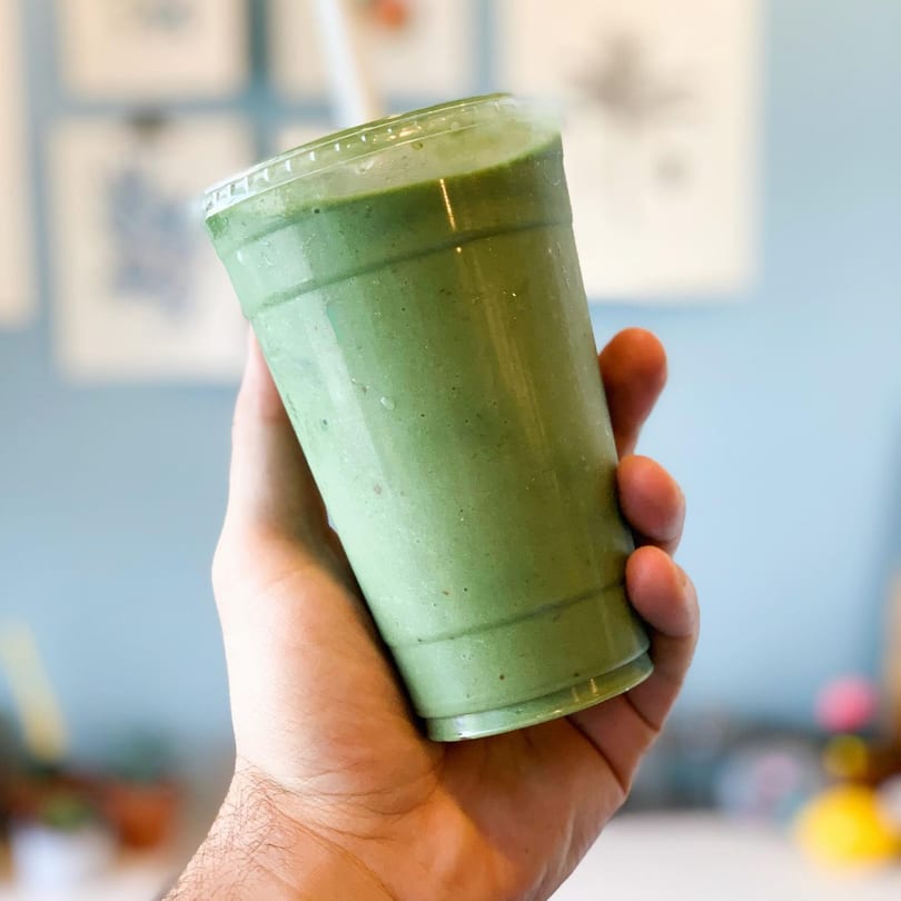 A hand holds up a green smoothie in a clear plastic cup from Juice Parlor NoHo.