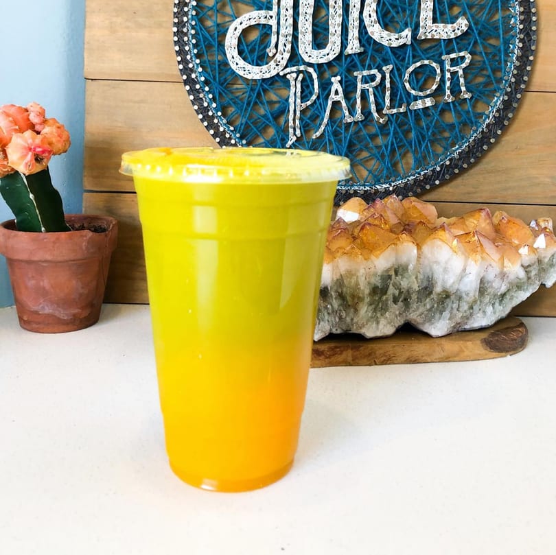 A vibrant yellow and orange layered juice in a clear cup, with a Juice Parlor sign in the background.